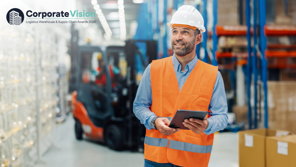 Worker in a warehouse wearing an orange safety vest and smiling holding a handheld device