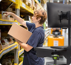 A strong woman is reaching up to place a box on a shelf in a warehouse, featuring ecommerce mobile computer cart