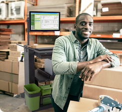 Man leaning on a stack of boxes in a warehouse, smiling at the camera, featuring ecommerce mobile computer cart