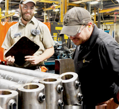 Two men, one holding a clipboard, and the other examining or working with metal pipes