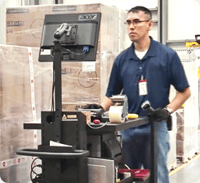 A worker operating a mobile workstation in a warehouse, with stacked pallets of boxes in the background.